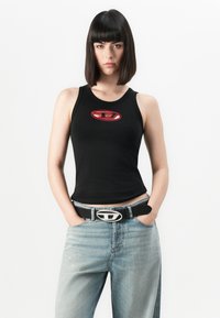 Junge Person mit schulterlangen, dunklen Haaren, die ein schwarzes, ärmelloses Top mit rotem Logo, hellblaue Jeans und einen schwarzen Gürtel mit silberner Schnalle trägt.