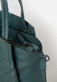 Primo piano di una borsa in pelle turchese che mostra le maniglie, le cuciture e un comparto interno con zip parzialmente visibile, dotato di una linguetta in pelle.