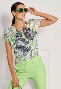 Multikleurige top met bloemen- en paisleypatronen, korte mouwen en een colnek, gecombineerd met groene broek. Opvallende groene zonnebril en sieraden.