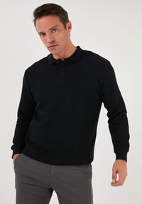 Polo de manga larga negro con textura acanalada, tapeta de tres botones y puños acanalados. Combinado con pantalones grises.
