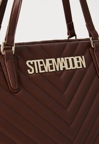 Ruda siuvinėta rankinė su tekstūruotu paviršiumi, su auksiniu "STEVE MADDEN" logotipu ir dviem petiniais diržais nešimui.