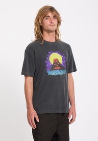 Homme aux cheveux blonds mi-longs portant un t-shirt noir avec un graphisme volcan coloré et un pantalon noir, debout devant un fond uni.