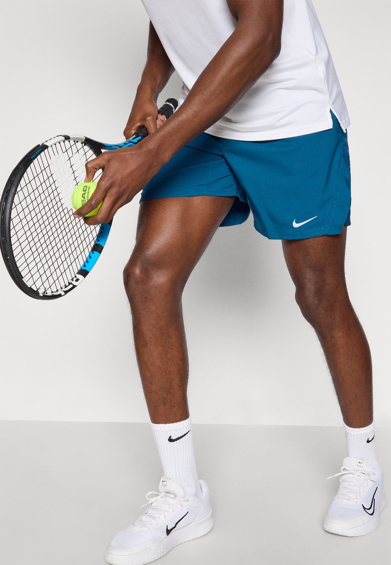 Blå sportshorts med Nike-logga, vit t-shirt, håller i en tennisracket och boll. Vita sneakers med svart Nike-logga.