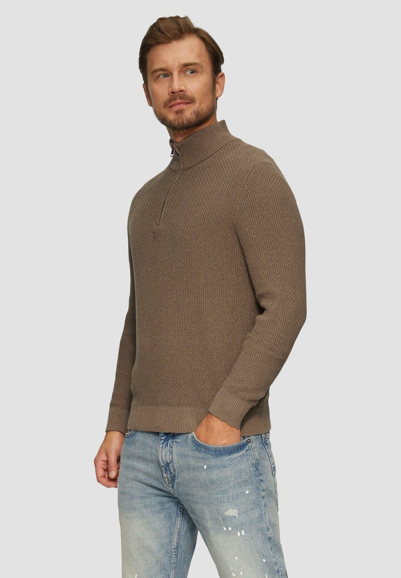 Brązowy sweter o teksturowanej powierzchni z wysokim kołnierzem i zamkiem, zestawiony z jasnoniebieskimi dżinsami w stylu vintage.