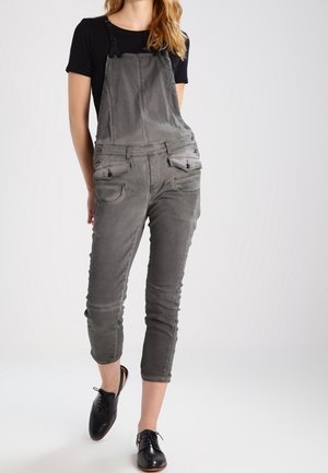 Femme portant une salopette grise raccourcie avec des poches avant et un t-shirt noir à manches courtes, associée à des chaussures noires à lacets, debout sur fond blanc.