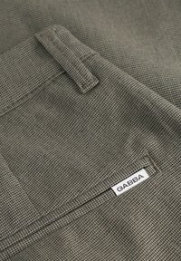 Tissu gris texturé avec un motif tissé, présentant une petite poche et une étiquette blanche avec "GABBA". Vue rapprochée du matériau et des détails de conception.