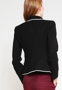 Svart skräddarsydd kavaj med en subtil textur, vit kant längs kragen och utsvängd nederdel, matchad med mönstrade röda byxor.