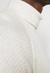 Krémový žebrovaný rolákový svetr s texturovanou tkaninou, který má vysoký límec a jemné vertikální vzory. Detail ramene.