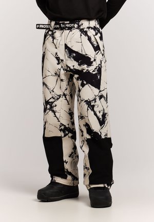Persona con pantalones de patrón abstracto en blanco y negro con remiendos negros en la parte inferior de las piernas y zapatos negros, de pie frente a un fondo liso.