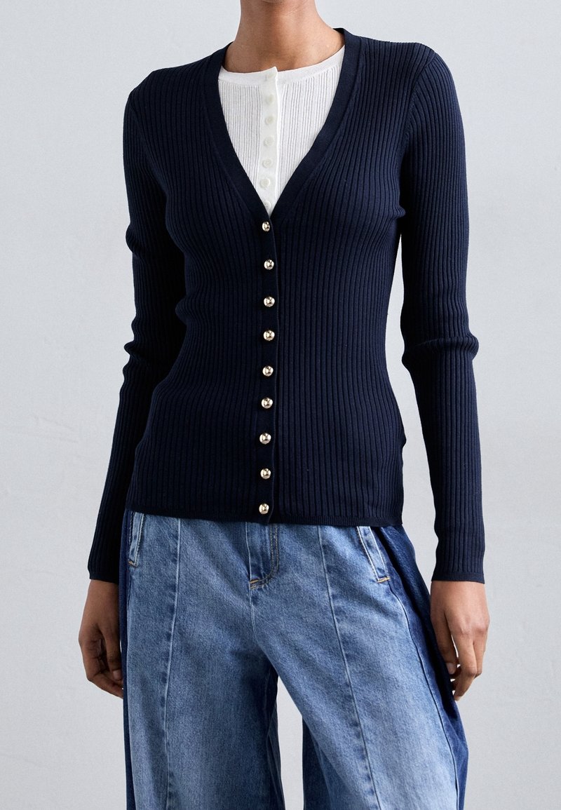 Femme portant un cardigan côtelé bleu marine avec des boutons argentés par-dessus un haut henley blanc et un jean bleu ample.