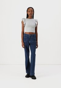 Siv cropped top z nabranimi rokavi, v kombinaciji z modrimi ravnimi hlačami. Model stoji na nevtralni podlagi, obut v črne čevlje.
