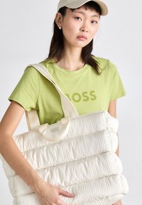 Cream-colored padded tote bag with vertical rows, light fabric, and dual flat handles. Model wears a green short-sleeve t-shirt.