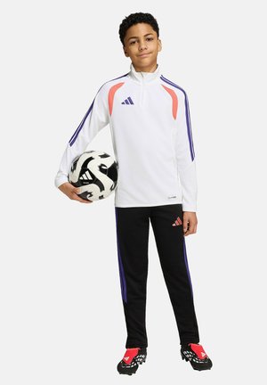 Ung dreng i hvid sportsjakke og sorte bukser holder en sort-hvid fodbold, iført fodboldstøvler, står mod en ensfarvet baggrund.