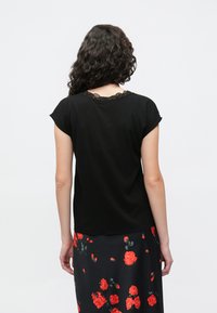 Svart topp med kappelignende ermer og en bølget blonderkant på utringningen, kombinert med et svart skjørt med røde roser. Stoffet ser glatt og mykt ut.