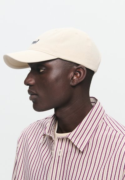 Vyras kremiško spalvos beisbolo kepurėje ir purpurinės bei baltos dryžuotos marškinėlių su mygtukais, žiūrintis į kairę prieš paprastą baltą fono.