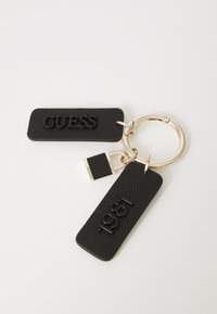 Czarny teksturowany brelok z tagami "GUESS" i "1981", małym czarnym i złotym urokiem w kształcie kłódki, przymocowany do złotego kółka na klucze.