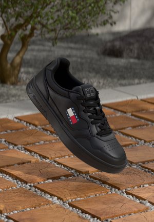 Czarny but sportowy Tommy Jeans z logo z boku i na języku, unoszący się nad drewnianymi płytkami i szarymi kamykami na zewnątrz.