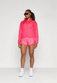 Chaqueta rosa ligera con cremallera, pantalones cortos rosas a juego y zapatillas blancas. El conjunto presenta un diseño deportivo y material transpirable.