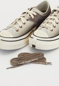 Zapatillas beige y marrones que presentan una combinación de texturas de tela, punta redonda, suela de goma blanca y cordones de marca. Incluye cordones marrones adicionales.