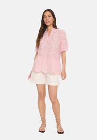 Blusa rosa in pizzo con maniche a sbuffo corte, orlo peplum e scollatura a V, abbinata a pantaloni corti beige chiaro.