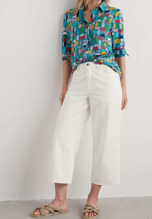 Femme portant un pantalon blanc large et court, une chemise à manches longues colorée et à motifs, et des sandales beiges en corde, se tenant contre un mur uni.