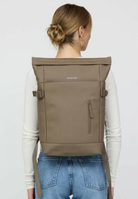 Brauner Rucksack mit Rollverschluss aus strapazierfähigem Material, seitlichen Riemen, einer vorderen Reißverschlusstasche und einem minimalistischen Logo.