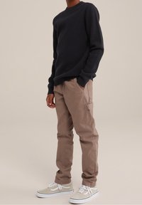 Hombre vestido con un suéter negro de manga larga, pantalones marrones con bolsillos laterales y zapatillas beige con cordones blancos. Manos en los bolsillos.