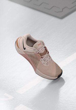 Hellrosa und beige Nike Laufschuh mit Mesh-Obermaterial und schwarzem Swoosh, schwebend über einem weißen Boden mit geometrischen Linien dargestellt.