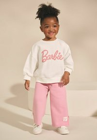 Weißes Sweatshirt mit "Barbie" in pinker Schrift, kombiniert mit rosa weit geschnittenen Sweatpants. Die Schuhe sind weiß und es gibt ein Etikett an der Hose.