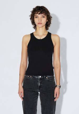 Jonge persoon met kort krullend haar, gekleed in een zwart mouwloos shirt, donkere spijkerbroek, zilveren ketting en armbanden, staand tegen een witte muur.