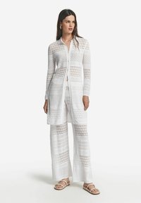 Vit, stickad överdragsoutfit som består av en lång, knappad cardigan och vida byxor med geometriska mönster och öppen vävning.