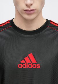 Czarna sportowa koszulka z czerwonymi trzema paskami na ramionach i czerwonym logo "adidas" na piersi. Gładka tekstura tkaniny.
