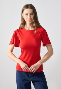 Top roșu cu mâneci scurte, cu un design neted, ajustat și cu mâneci evazate, fabricat dintr-un material ușor. Asortat cu blugi negri.