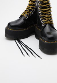 Bottes de combat plates noires avec des lacets épais jaunes et noirs, des coutures jaunes et des lacets noirs extra longs et lâches sur fond blanc.
