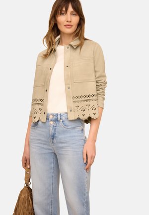 Femme portant une veste beige avec un ourlet festonné découpé au laser, un haut blanc, un jean bleu clair et tenant un sac à main marron à franges à côté d'elle.