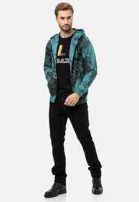 Grün-blauer Reißverschluss-Hoodie mit abstrakten Mustern, getragen über einem schwarzen T-Shirt mit einem bunten Logo, kombiniert mit schwarzen Hosen und Stiefeln.