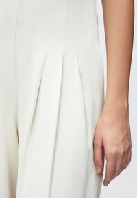 Material crem plisat pentru salopetă, cu o textură fină. Prim-planul arată detaliile pliurilor ascuțite și o croială lejeră, cu o mână sprijinindu-se pe lateral.