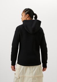 Melnas kapuces sporta jaka ar brīvu piegriezumu, kas aprīkota ar kengura kabatu un ribinātām piedurknēm, valkāta pār gaišām krāsainām biksēm.