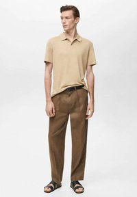 Khaki polo triko, hnědé kalhoty, černé sandály. Polo má límec a krátké rukávy. Kalhoty mají volný střih a hladkou texturu.