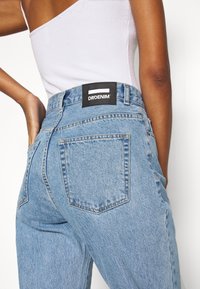 Světle modré džíny s vysokým pasem a výrazným designem zadní kapsy. Na pásce je černá etiketa s bílým textem.
