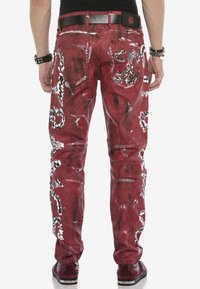 Rote Jeans aus Denim mit Farbspritzern und abstrakten Mustern; mit Taschen und geradem Beinschnitt. Schwarzes Gürtel-Detail sichtbar.