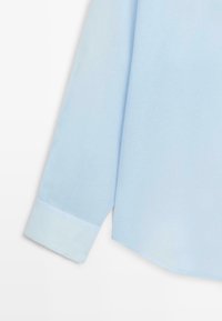 Lichtblauw overhemd met lange mouwen en een knoopmanchet, zijaanzicht tegen een effen witte achtergrond.