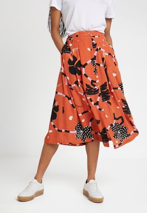 Femme portant un t-shirt blanc, une jupe midi orange avec des motifs floraux abstraits noirs, roses et blancs, et des baskets blanches avec des semelles en gomme.