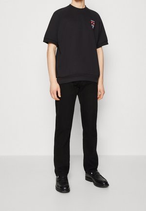 Schwarzes Baumwoll-T-Shirt in Übergröße mit kurzen Ärmeln und geripptem Rundhalsausschnitt. Mit einem grafischen Design auf der Brust. Kombiniert mit schwarzen Hosen und Schuhen.