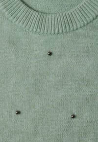Pull tricoté vert clair avec col côtelé et trois petits clous métalliques ronds disposés en motif triangulaire à l'avant.