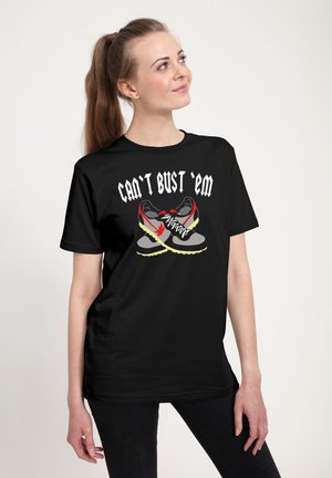 Jonge vrouw in een zwart T-shirt met een afbeelding van gekruiste sneakers en de tekst "Can't Bust 'Em", kijkt omhoog en glimlacht lichtjes.