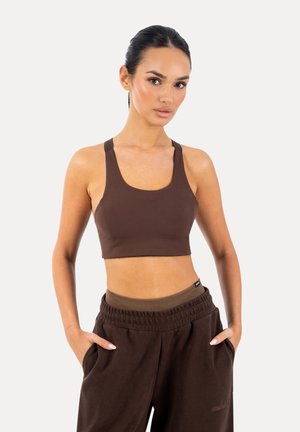 Mujer con un sujetador deportivo marrón y pantalones deportivos de cintura alta a juego, con las manos en los bolsillos, de pie frente a un fondo blanco liso.