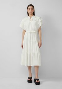 Witte gelaagde jurk met ruffle-mouwen, elastische tailleband en een keyhole-halslijn. Gekoord met zwarte platform sandaaltjes. Effen, lichtgewicht stof.