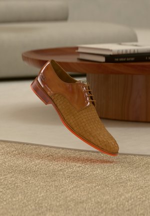 Chaussure habillée en cuir marron à texture tissée, flottant au-dessus d'un tapis beige dans un salon moderne avec une table en bois et des livres.