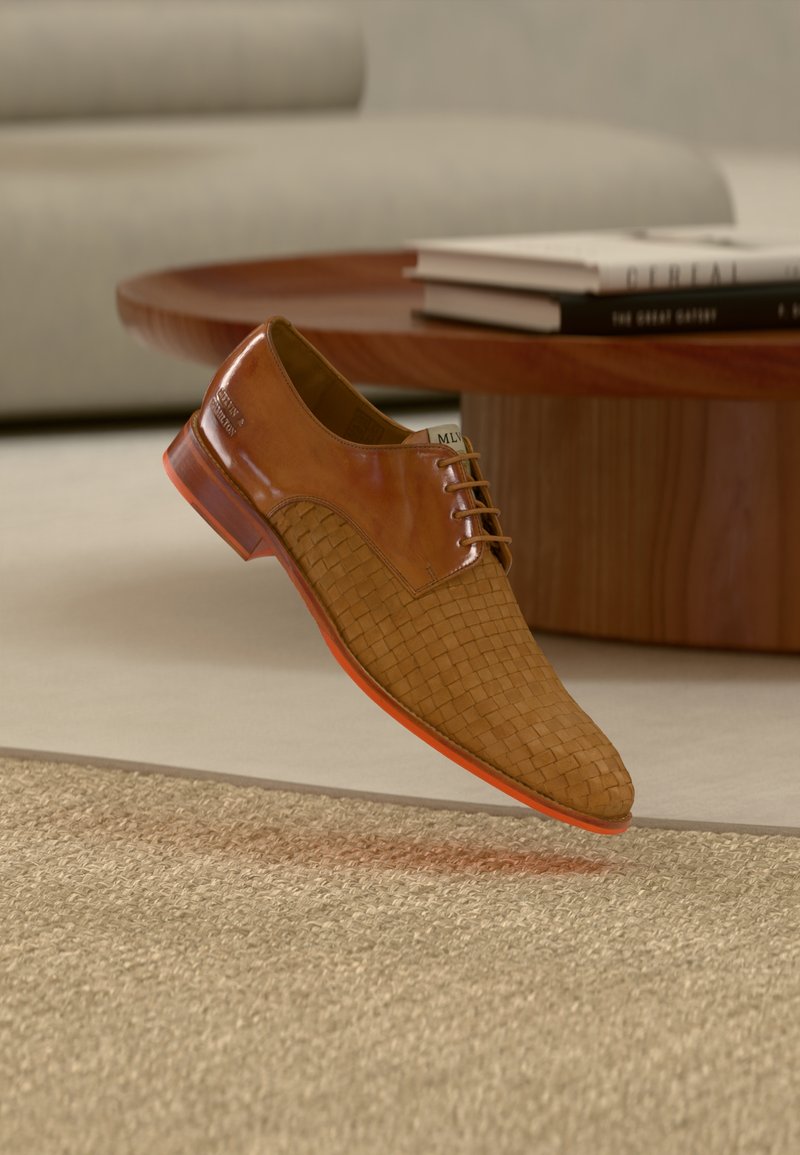 Chaussure habillée en cuir marron à texture tissée, flottant au-dessus d'un tapis beige dans un salon moderne avec une table en bois et des livres.